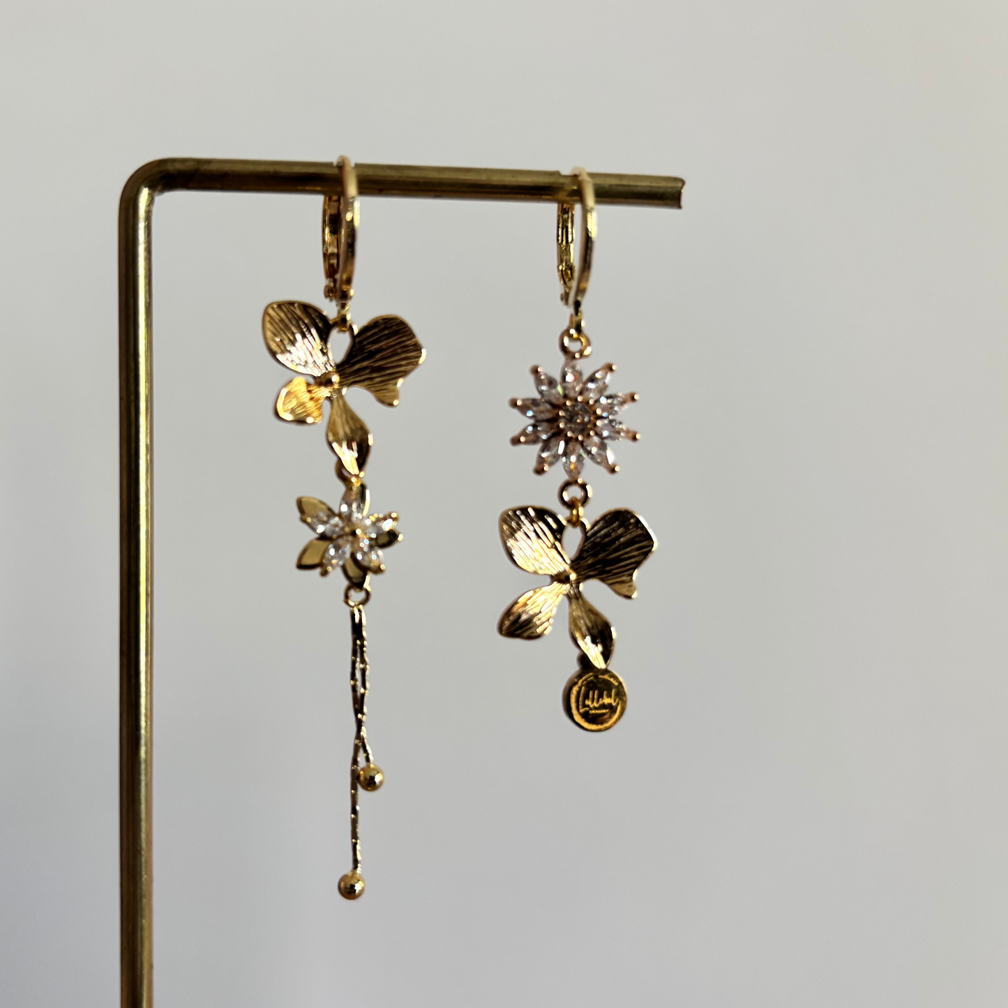 Asymmetrische goudkleurige oorbellen Roselien aan een rekje. Links een gouden bloem met een zirkonia ster en lang hangertje; rechts een zirkonia bloem met een gouden bloem en merkbedeltje.