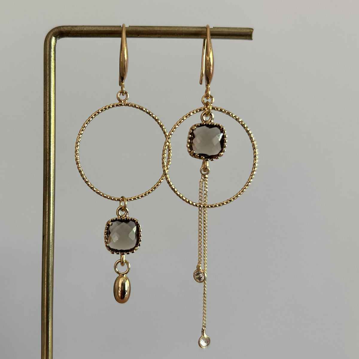 Goudkleurige asymmetrische oorbellen Cara met bewerkte ronde ringen, vierkante terrakleurige kristallen en fijne kettinkjes met zirkonia.