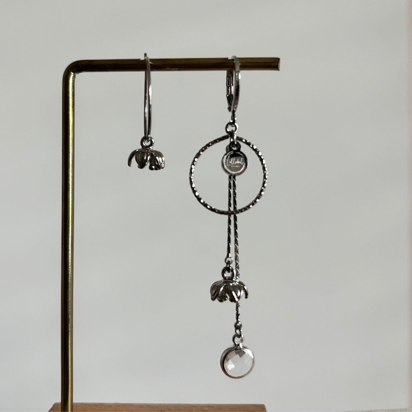 Asymmetrische zilverkleurige oorbellen Daphne mismatch aan een rekje. Links een korte hanger met een klein tulpvormig bloemetje; rechts een grote bewerkte cirkel met lange kettinkjes, een bloemetje en een ronde heldere kristalsteen.