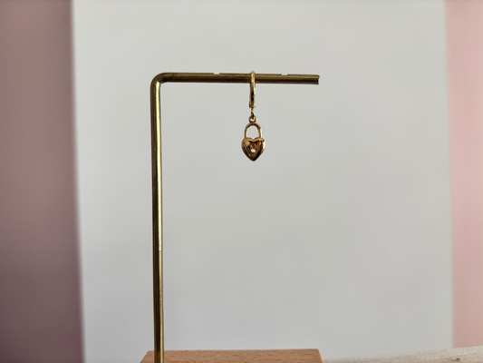 Een goudkleurig klapoorringetje met een hangend hartvormig slotje als bedel, gepresenteerd op een gouden displaystandaard tegen een wit-roze achtergrond.