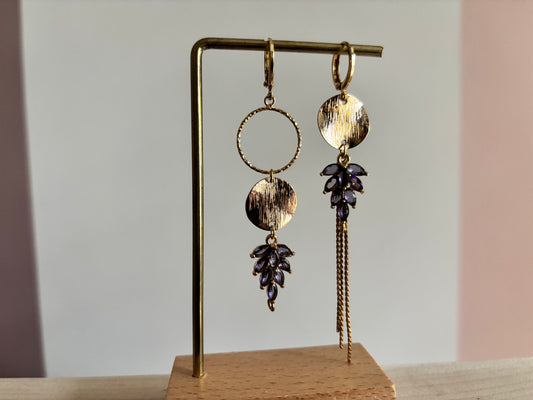 Gouden asymmetrische oorbellen Fabienne met paarse steentjes op een standaard.