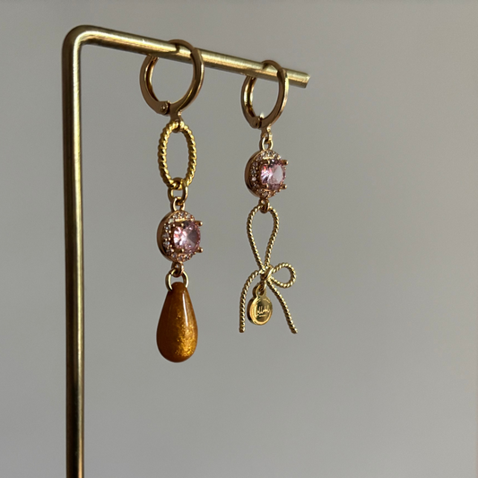Gouden asymmetrische oorbellen Harper met roze kristal, strikje en okerkleurige druppel op een standaard.