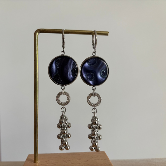 Zilverkleurige oorbellen Joke aan een rekje. Model met een ronde donkerblauwe polaris steen, een klein strass-ringetje en een hangend trosje van zilveren bolletjes.