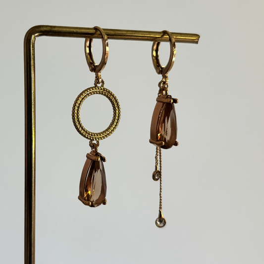 Set asymmetrische goudkleurige oorbellen aan een display. Links een opengewerkte ronde cirkel met daaronder een hangende, rookbruine facetgeslepen druppelsteen. Rechts dezelfde bruine druppelsteen met daaronder twee fijne goudkleurige kettingkjes met kleine steentjes.