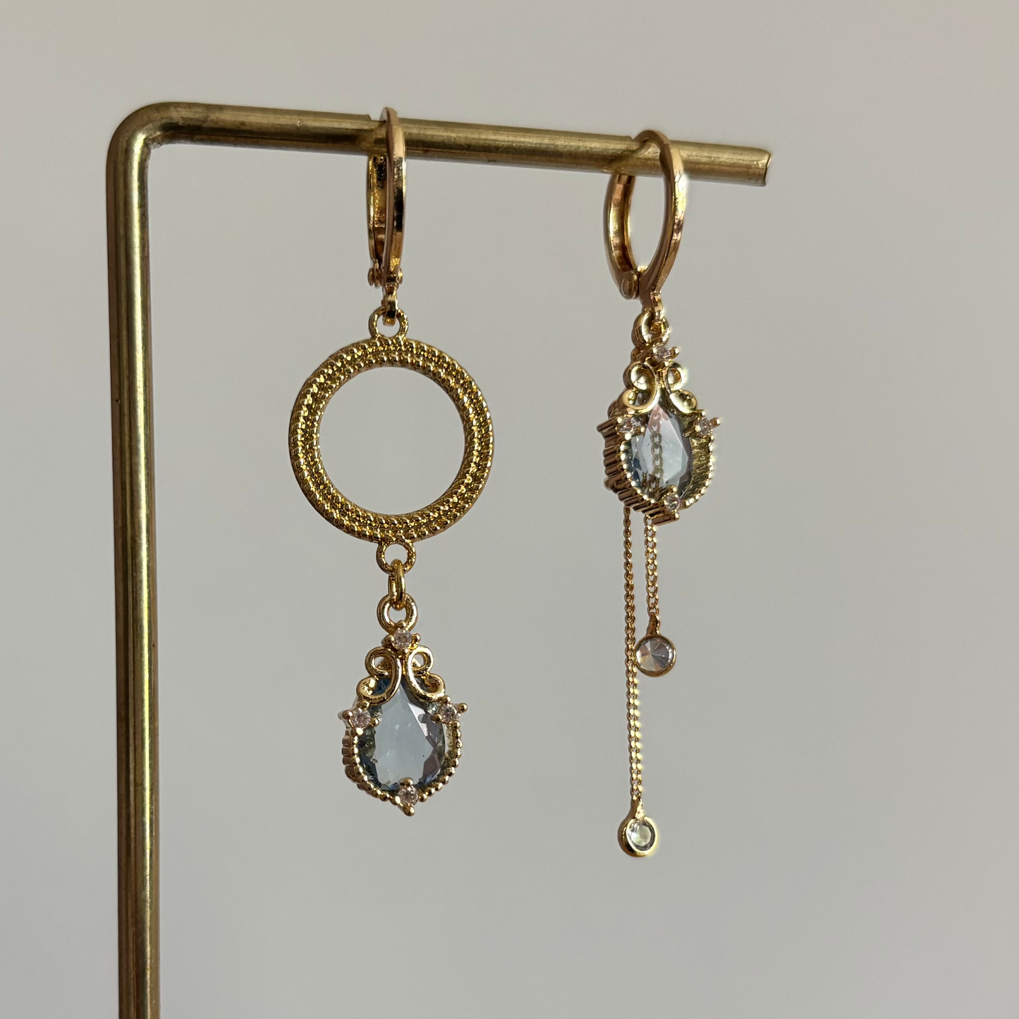 Set asymmetrische goudkleurige oorbellen aan een display. Links een opengewerkte ronde cirkel met daaronder een hangende, lichtblauwe facetgeslepen druppelsteen. Rechts dezelfde blauwe druppelsteen met daaronder twee fijne goudkleurige kettingkjes met kleine steentjes.