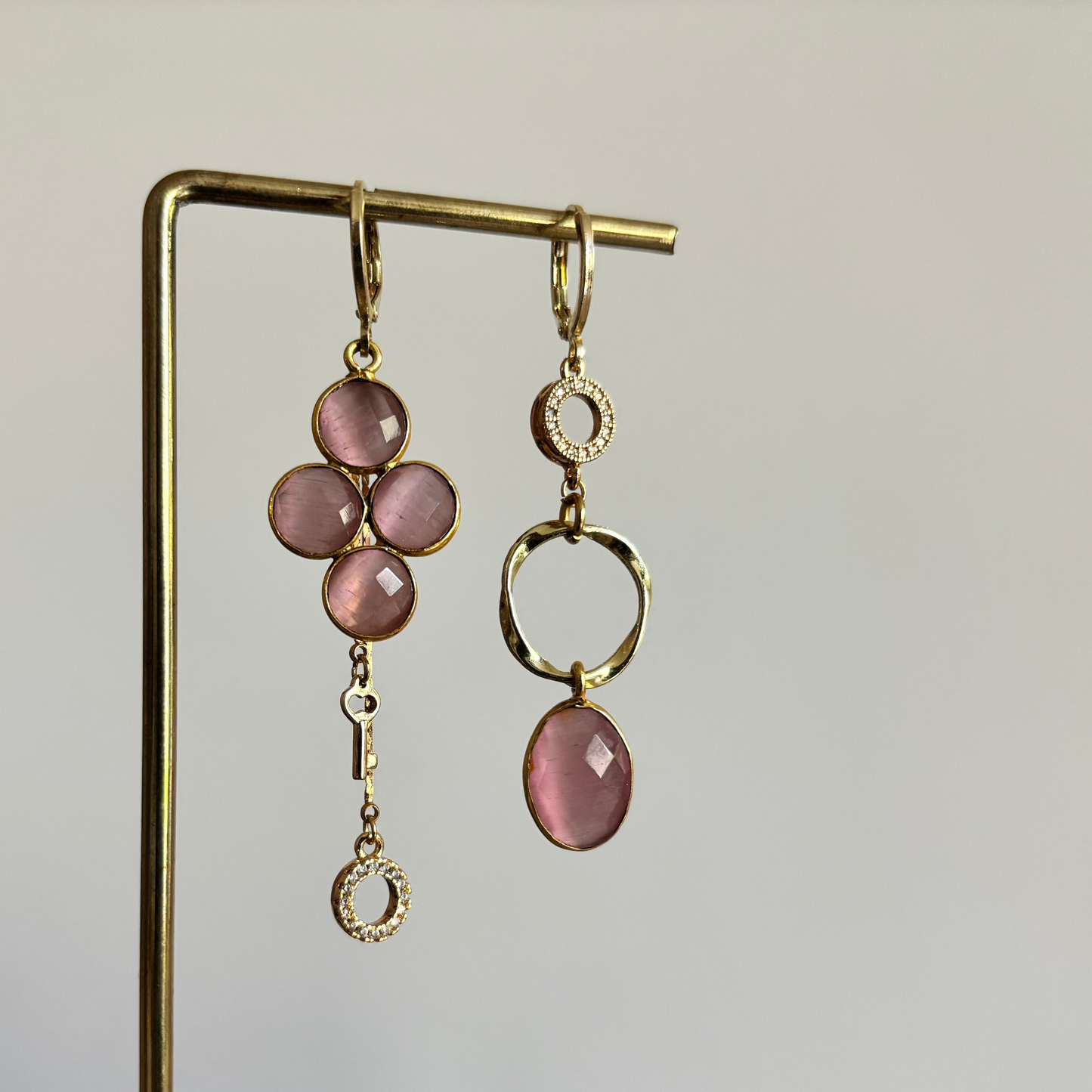 Asymmetrisch oorbellenpaar ‘Lola’: linker oorbel met zirkonia steentje, mini twisted ring en roze glazen bedel; rechter oorbel met taupekleurige steen, kwastje met sleuteltje en zirkonia steentje. Verguld in 14k goud, minimalistisch ontwerp met subtiele details.