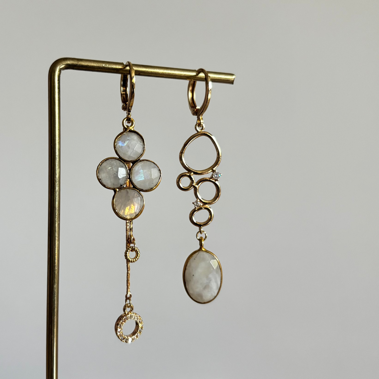 Asymmetrische goudkleurige oorbellen Lola'tje Maansteen aan een rekje. Links een klavervorm van vier maanstenen met een lang hangertje; rechts een speelse schakel van organische ringen met een ovale maansteen.
