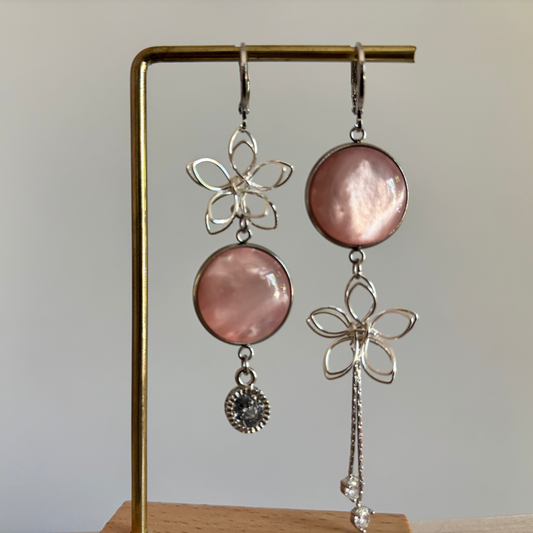 Asymmetrische zilveren oorbellen van Lellebel op een gouden standaard. De oorbellen bestaan uit een combinatie van zilveren draadbloemen, ronde zalmroze glansbedels, een klein zirkonia steentje aan de ene kant en een langer zilveren kettinkje aan de andere kant.