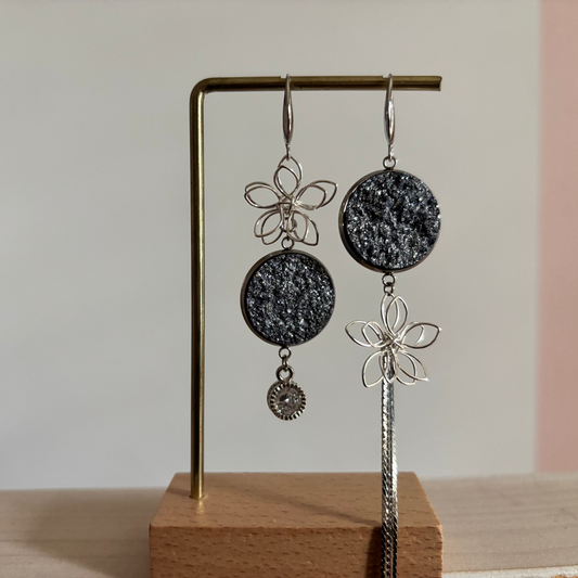 Asymmetrische zilverkleurige oorbellen Lou aan een rekje. Links een open zilveren draadbloem met een ronde lichtblauwe glittersteen en een ronde zirkonia hanger; rechts een lichtblauweglittersteen met een draadbloem en een lang kettinkje.