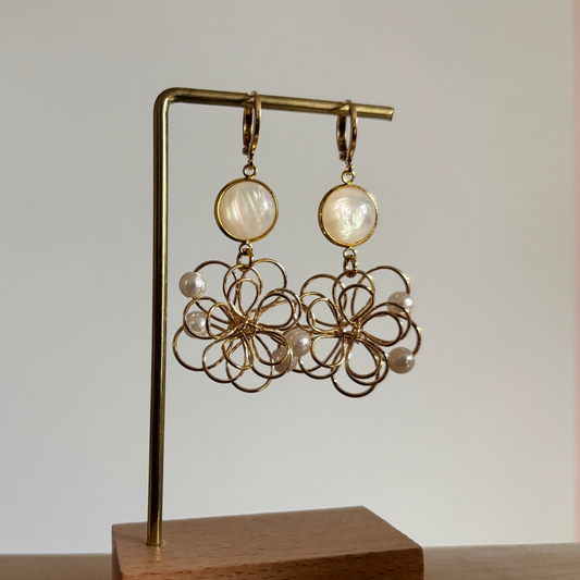 Symmetrische goudkleurige oorbellen 'Louise Parelmoer' op een houten display. De oorbellen bestaan uit een ronde, glanzende witte parelmoersteen en een grote opengewerkte bloemhanger van goudkleurig draad met kleine witte pareltjes.