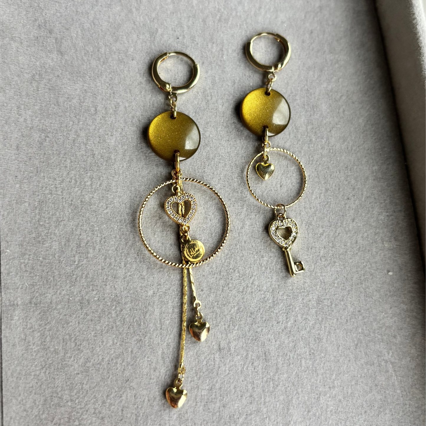 Asymmetrische goudkleurige oorbellen Nienke in een grijs sieradendoosje, met ronde olijfgroene stenen, fijne gedraaide ringen, een zirkonia hart, kleine hartjesbedels en een sleuteltje.