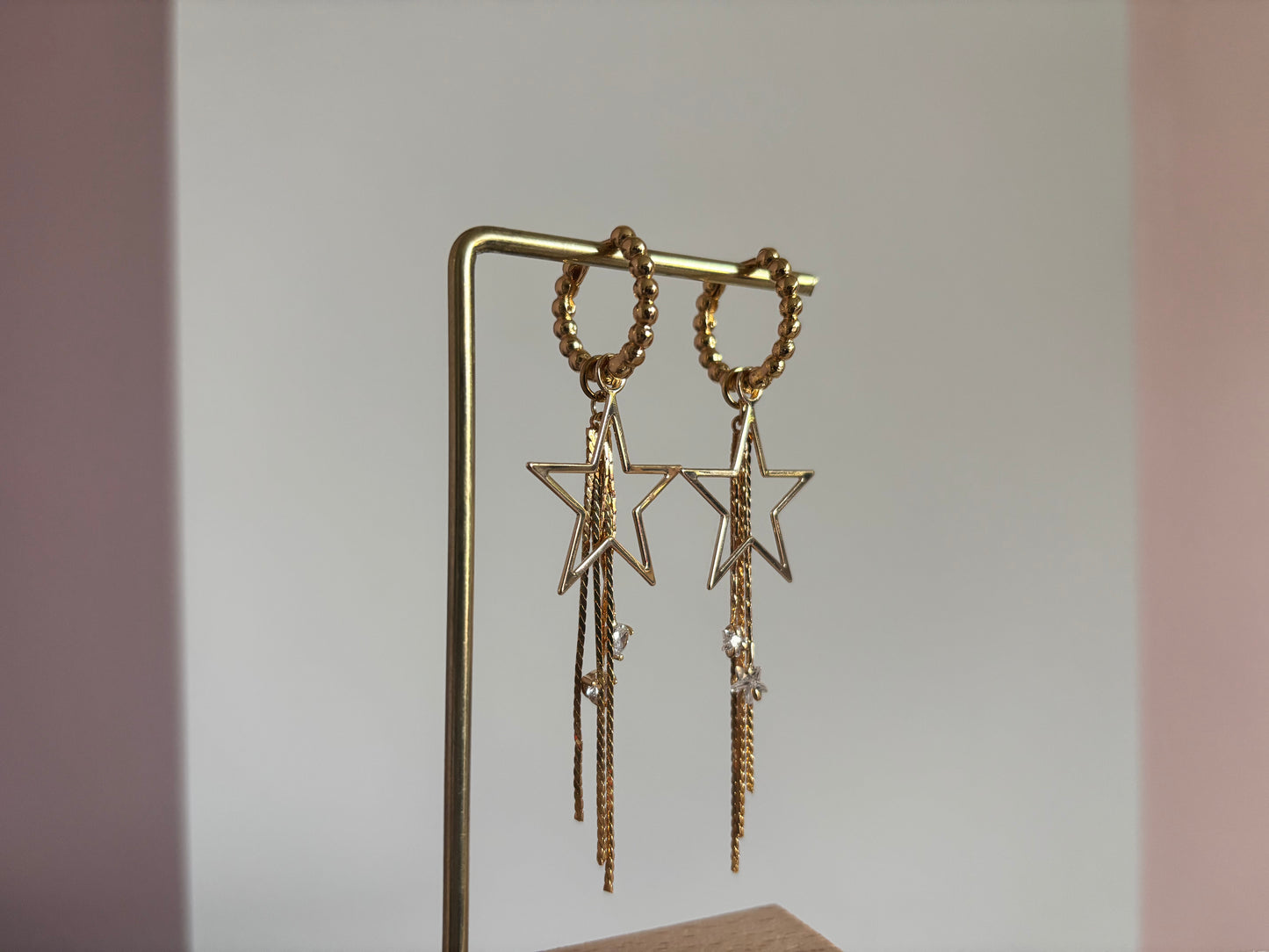 14k vergulde oorbellen 'Sterre' van Lellebel op een standaard. De oorbellen hebben een goudkleurige ster met daaraan hangende fijne kettinkjes en kleine glanzende zirkonia steentjes.