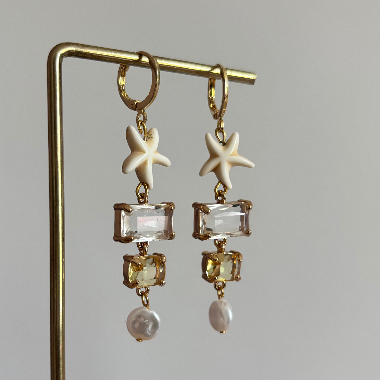Symmetrische goudkleurige oorbellen Summer aan een rekje, opgebouwd uit een witte zeester, een rechthoekig helder kristal, een geel kristal en een zoetwaterparel.