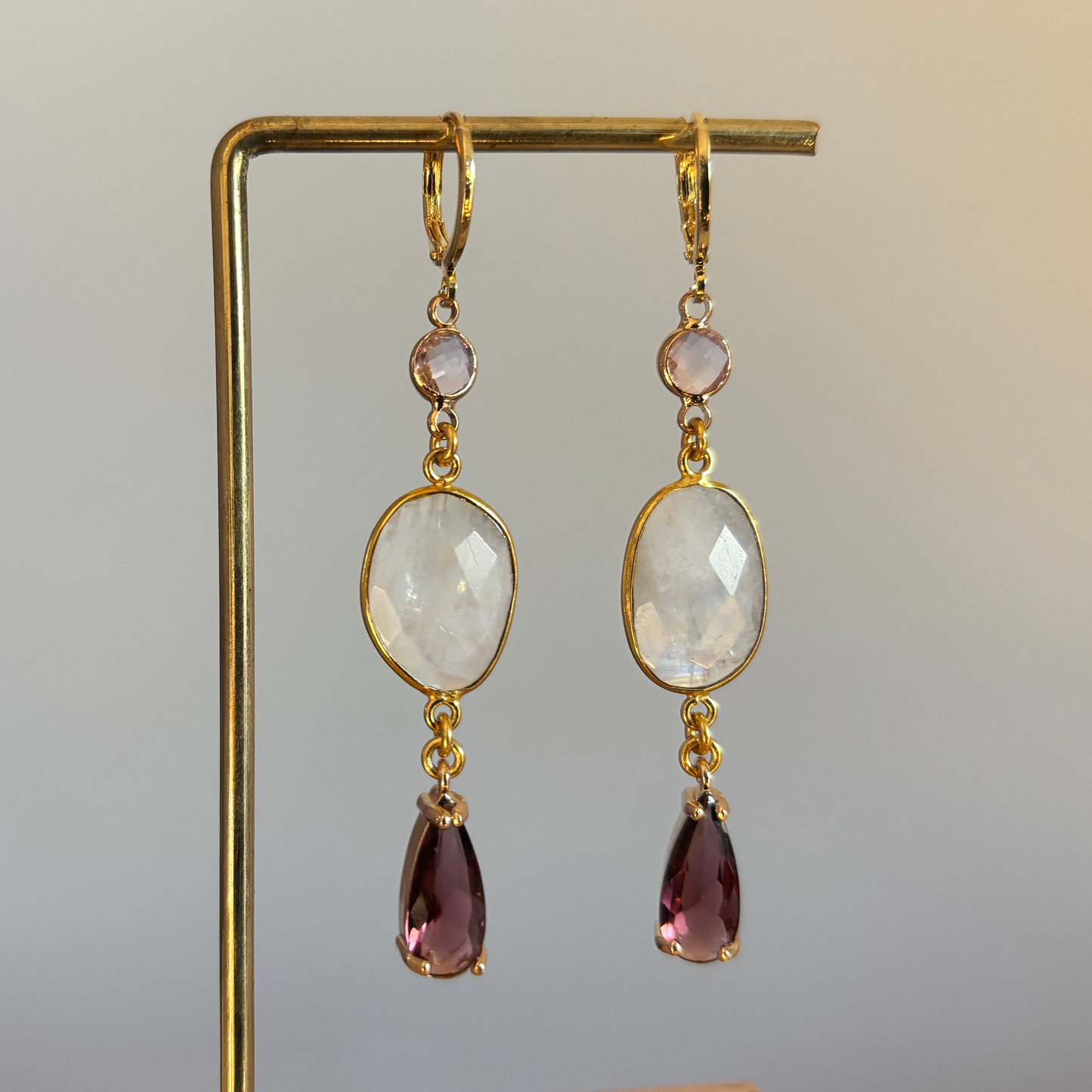 "Lange gouden oorbellen met een kleine lichtroze steen, een witte ovale steen en een dieppaarse facetsteen hanger."