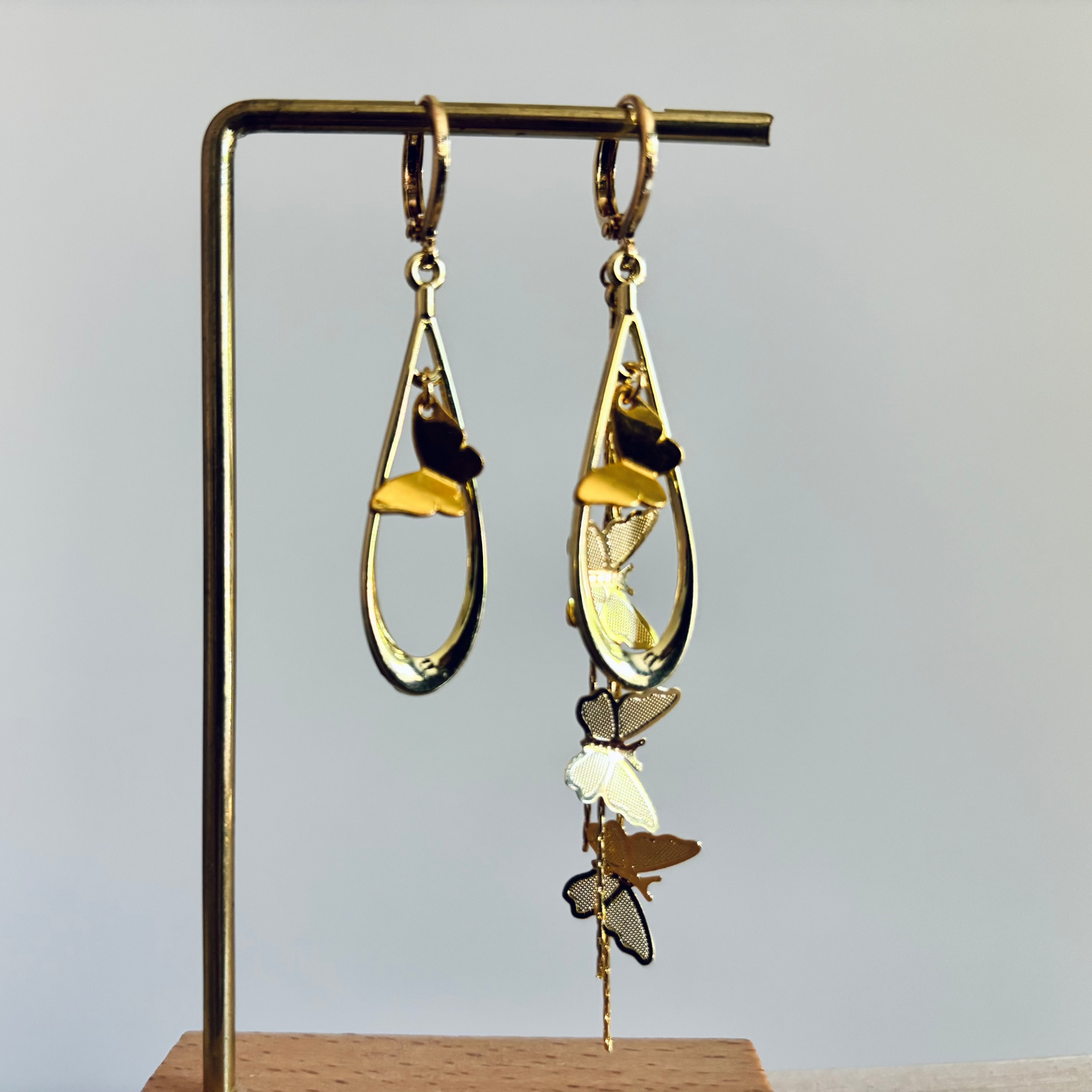 Asymmetrische gouden oorbellen van Lellebel op een standaard. Beide oorbellen hebben een open, druppelvormige hanger met daarin een klein gouden vlindertje. De rechteroorbel is extra lang door een sierlijke, verticale streng met meerdere gedetailleerde gouden vlinders die naar beneden hangen.