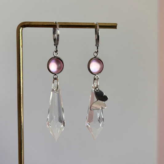 Asymmetrische zilverkleurige oorbellen genaamd 'Vlinder Zilver' op een display. Beiden hebben een ronde roze steen en een grote heldere acrylkristal. Rechts met een zilveren vlinderbedel