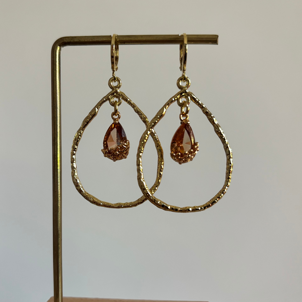 "Gouden symmetrische oorhangers Soraya Champagne met een grote bewerkte druppelvormige ring en een champagnekleurige kristallen druppelsteen daarin hangend."
