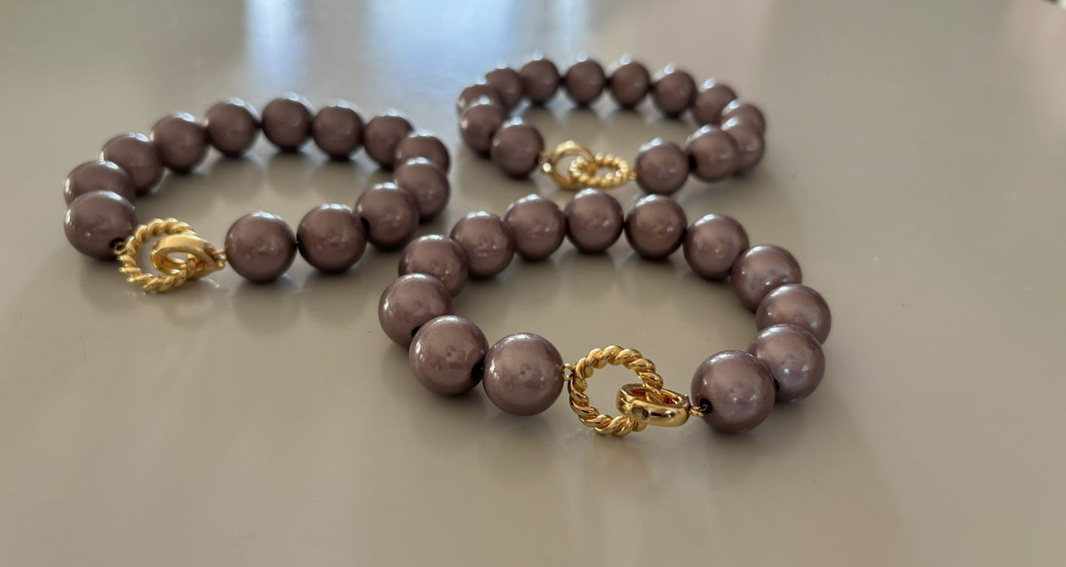Een kralenarmband genaamd Armband 3D Goud met ronde, taupe kralen, De armband heeft in het midden een opvallende goudkleurige sluiting bestaande uit een gladde ring en een gedraaid goudkleurig ringetje.