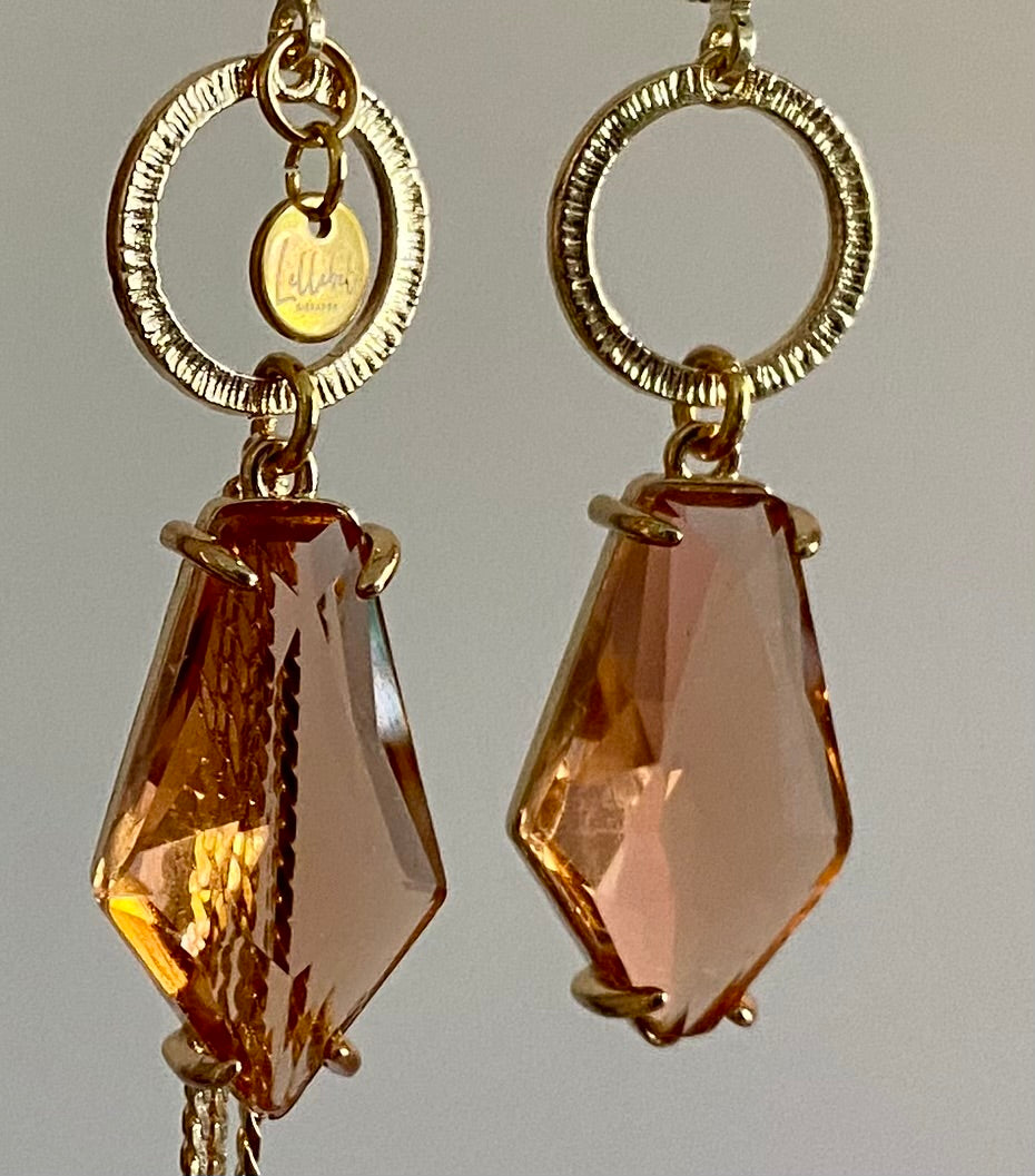 Close-up van Asymmetrische oorbellen met gehamerde gouden ring, klein merk logo, roest oranje glasbedel in de vorm van een hexagon en gouden streng 