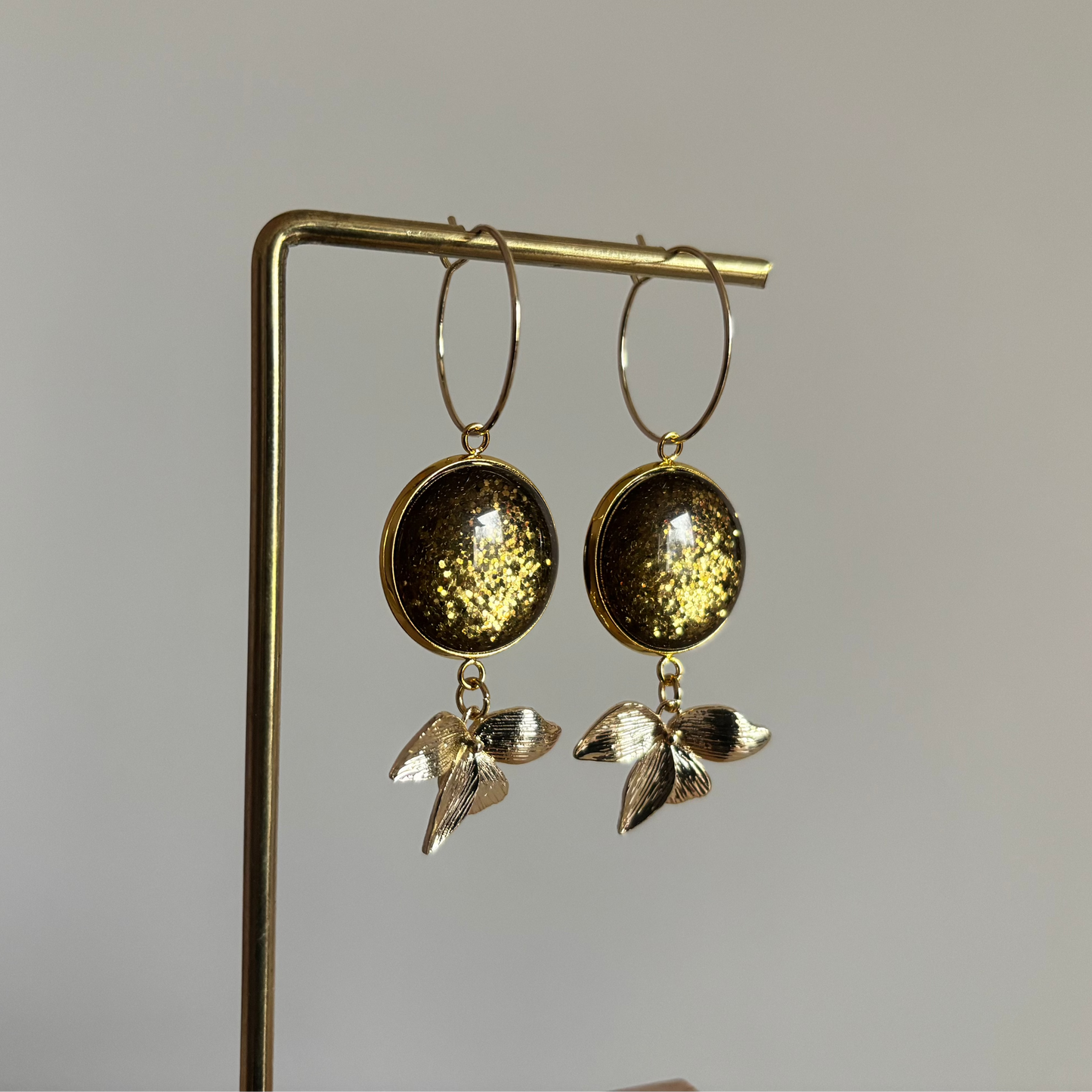 Gouden oorbellen met een 14k vergulde ring, een klein bladaccent en een ronde, groene glitter cabochon tegen een lichte achtergrond.