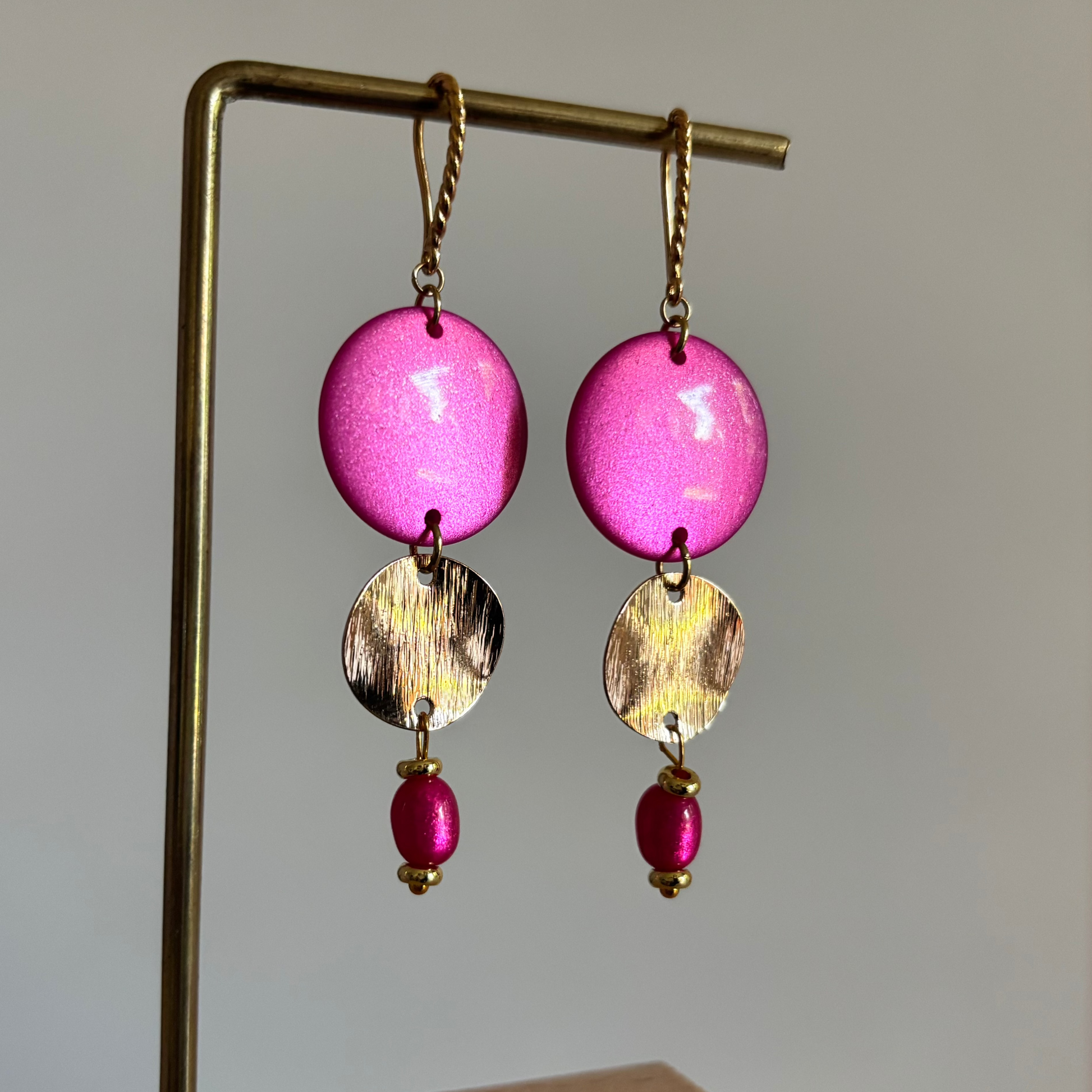 Symmetrische goudkleurige oorbellen Amanda aan een speels gedraaid oorhaakje, opgebouwd uit een grote, ronde roze polaris kraal, een gehamerd goudkleurig muntje en een klein roze kraaltje onderaan.