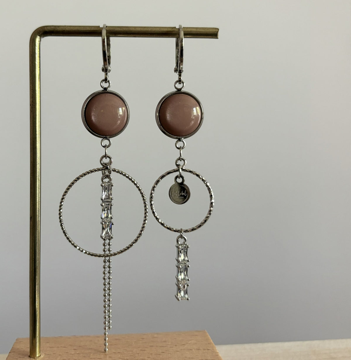 Twee verschillende zilveren oorbellen Anne op een standaard: beide voorzien van een ronde oudroze steen en ring, waarvan de ene een hangend kettinkje met strass heeft en de andere een bedel en onderhangende strass-steentjes.