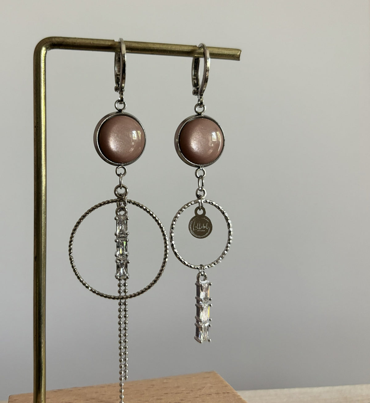 Twee verschillende zilveren oorbellen Anne op een standaard: beide voorzien van een ronde oudroze steen en ring, waarvan de ene een hangend kettinkje met strass heeft en de andere een bedel en onderhangende strass-steentjes.