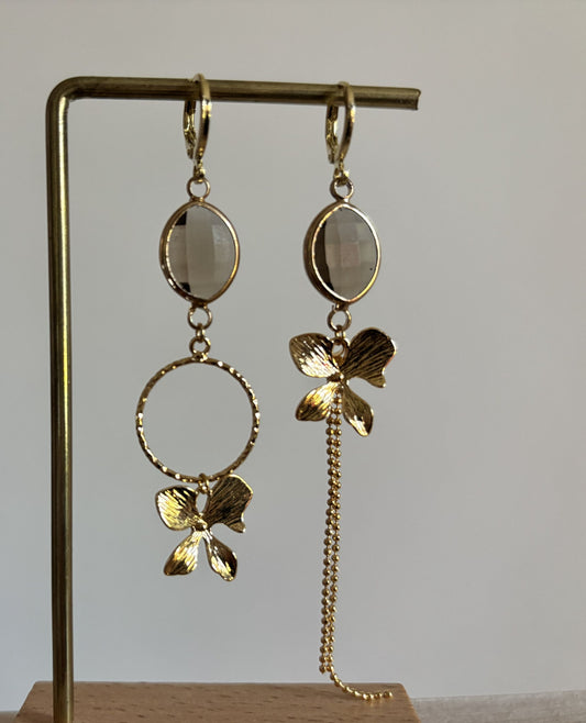 Goudkleurige asymmetrische oorbellen Annick Taupe met ovale grijze glasstenen, een bewerkte ring, gouden bloem met structuur en een fijn kettinkje.