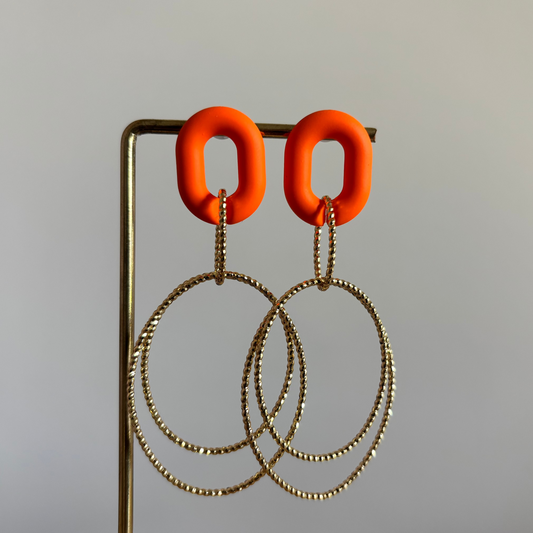 Een paar symmetrische Lellebel oorbellen genaamd Ariane, getoond op een gouden standaard tegen een neutrale achtergrond. De oorbellen hebben een matte oranje polymeer oorsteker met daaronder twee forse, in elkaar hangende 14k vergulde gouden hoops. De totale lengte is ongeveer 7 centimeter