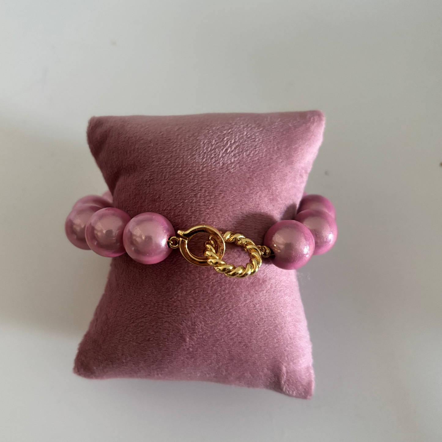 Een kralenarmband genaamd Armband 3D Goud met ronde, parelmoerroze kralen, gepresenteerd op een roze fluwelen kussentje. De armband heeft in het midden een opvallende goudkleurige sluiting bestaande uit een gladde ring en een gedraaid goudkleurig ringetje.