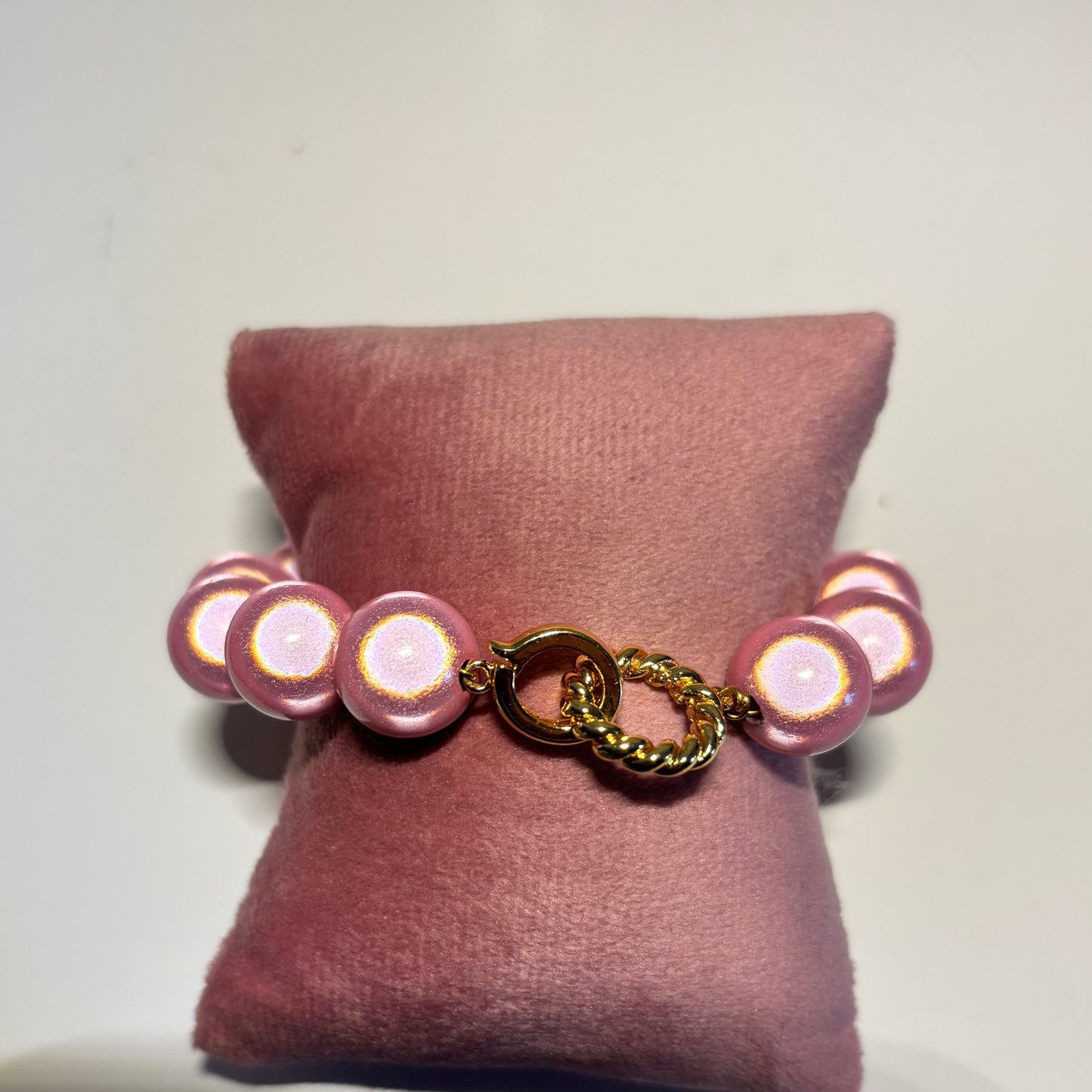 Een kralenarmband genaamd Armband 3D Goud met ronde, parelmoerroze kralen, gepresenteerd op een roze fluwelen kussentje. De armband heeft in het midden een opvallende goudkleurige sluiting bestaande uit een gladde ring en een gedraaid goudkleurig ringetje.