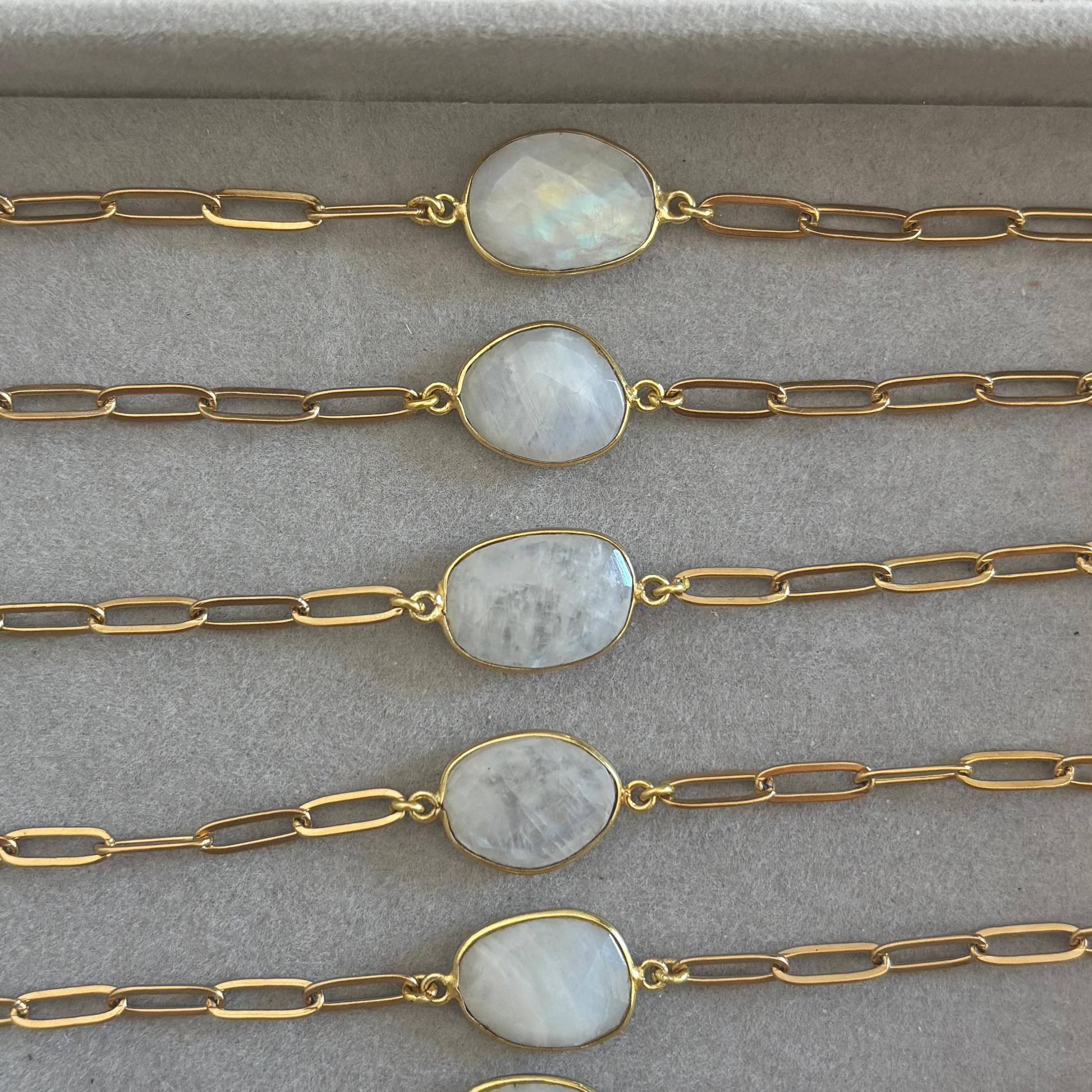 Zes goudkleurige Ada schakelarmbanden op een rij in een grijs sieradendoosje, elk voorzien van een grove langwerpige schakelketting en een centrale, ovale halfedelsteen met een wit-grijze glans.