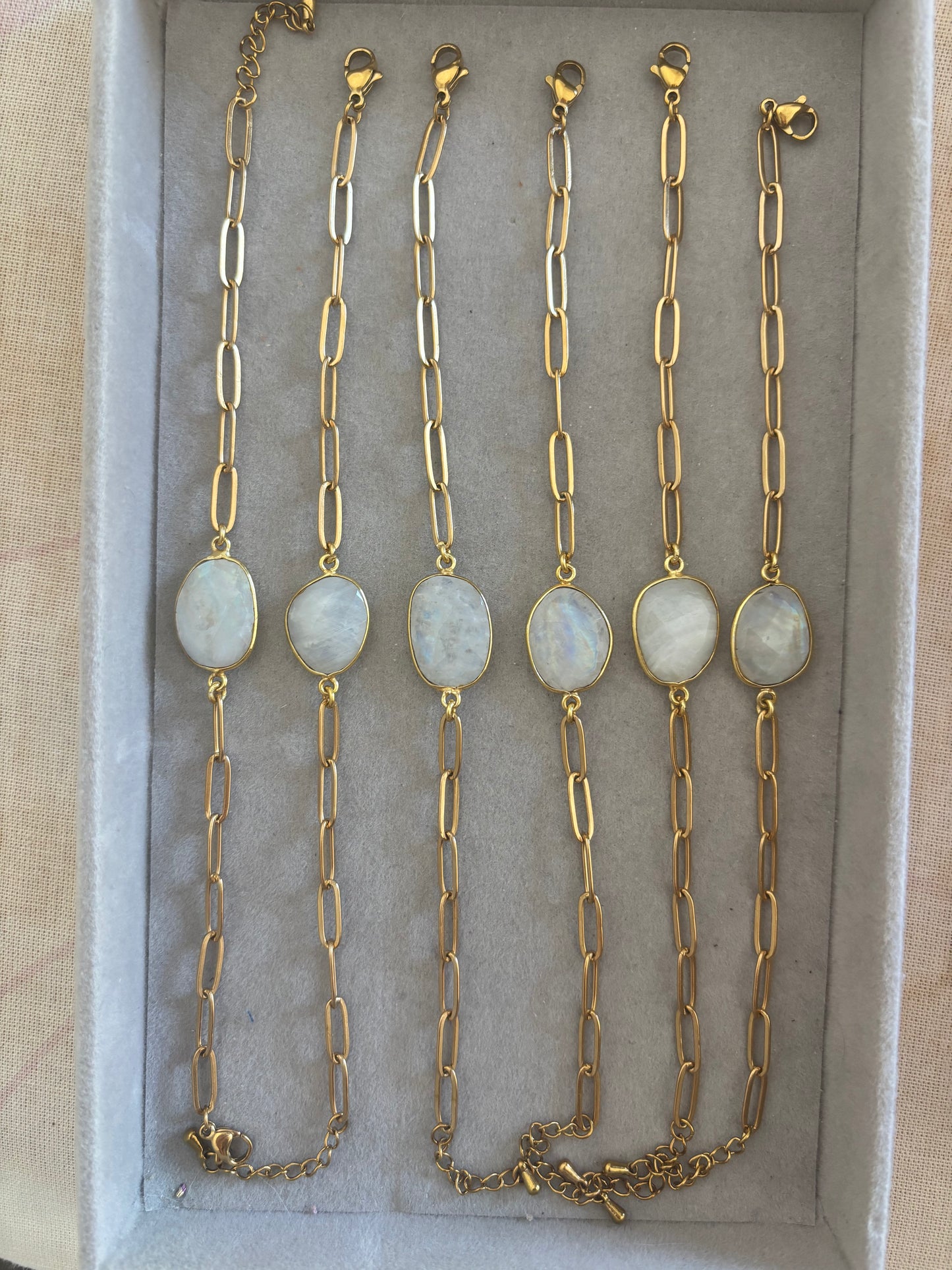 Zes goudkleurige Ada schakelarmbanden op een rij in een grijs sieradendoosje, elk voorzien van een grove langwerpige schakelketting en een centrale, ovale halfedelsteen met een wit-grijze glans.
