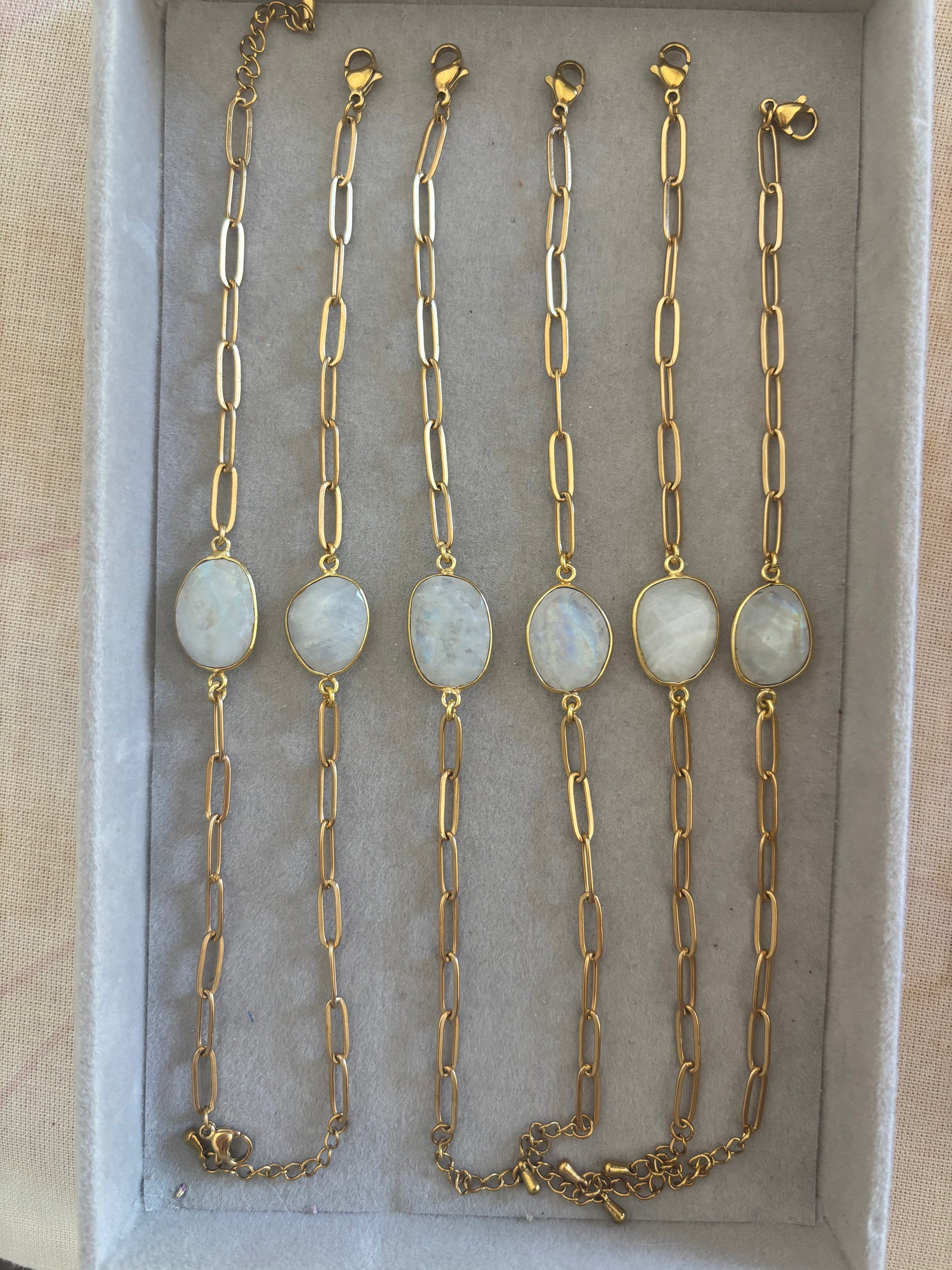 Zes goudkleurige Ada schakelarmbanden op een rij in een grijs sieradendoosje, elk voorzien van een grove langwerpige schakelketting en een centrale, ovale halfedelsteen met een wit-grijze glans.