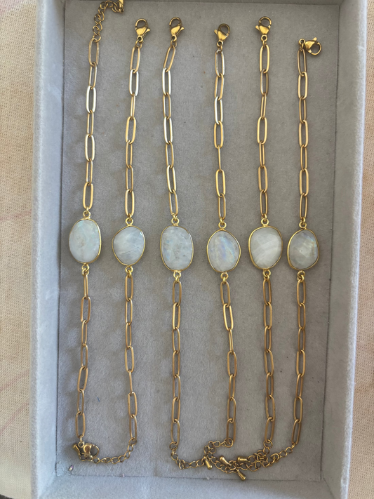 Zes goudkleurige Ada schakelarmbanden op een rij in een grijs sieradendoosje, elk voorzien van een grove langwerpige schakelketting en een centrale, ovale halfedelsteen met een wit-grijze glans.