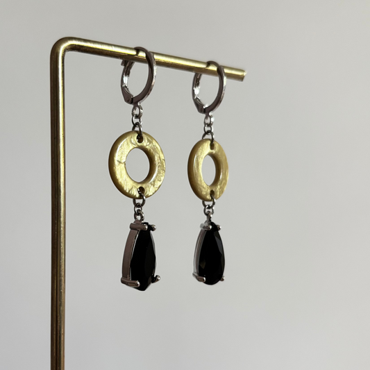 Zilverkleurige oorbellen Barbara aan een rekje. Model met een platte, ronde olijfgeel-groene ring en een hangende, facetgeslepen zwarte druppelsteen in een zilveren zetting.