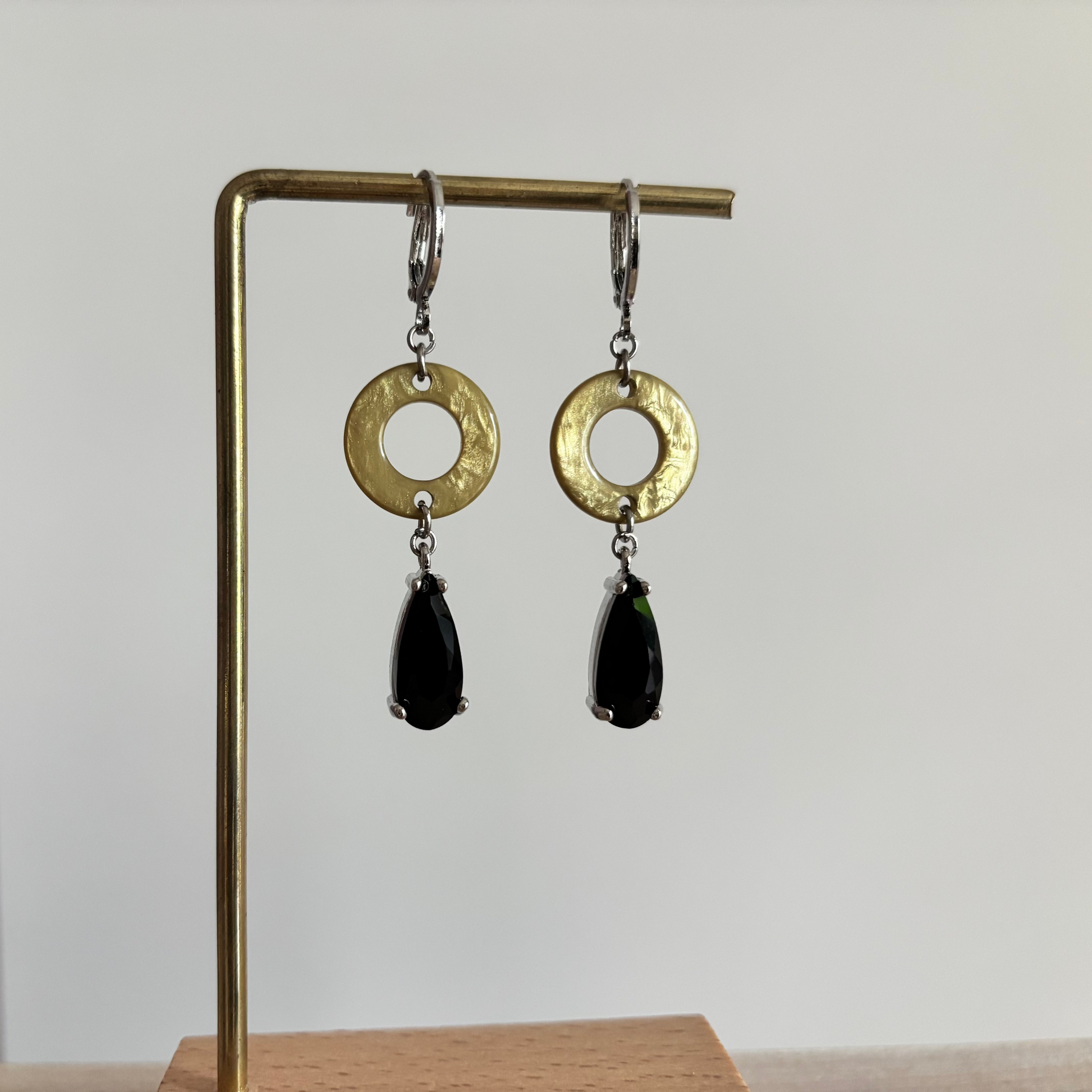 Zilverkleurige oorbellen Barbara aan een rekje. Model met een platte, ronde olijfgeel-groene ring en een hangende, facetgeslepen zwarte druppelsteen in een zilveren zetting.