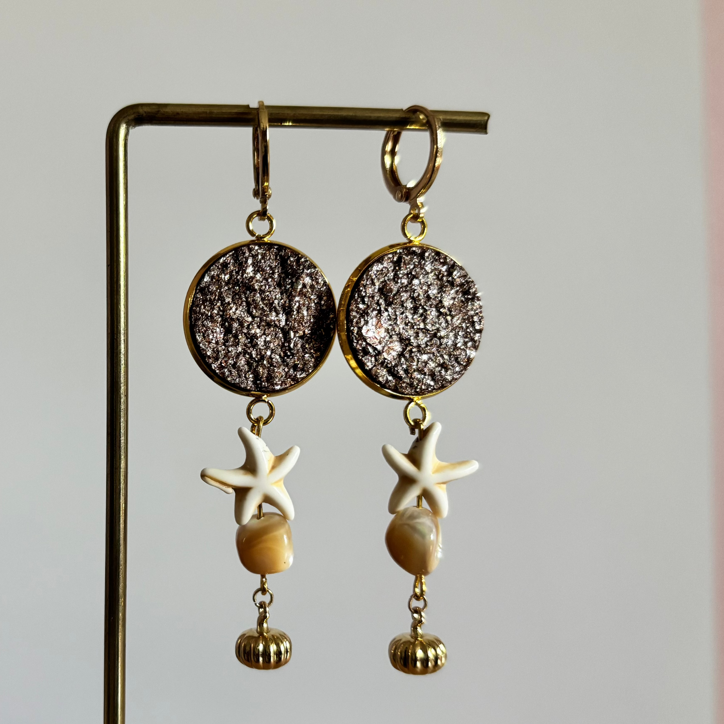 Symmetrische goudkleurige oorbellen Beachy aan een rekje, opgebouwd uit een ronde goudbruine glittersteen, een witte zeester, een beige schelpje en een geribbeld gouden kraaltje.