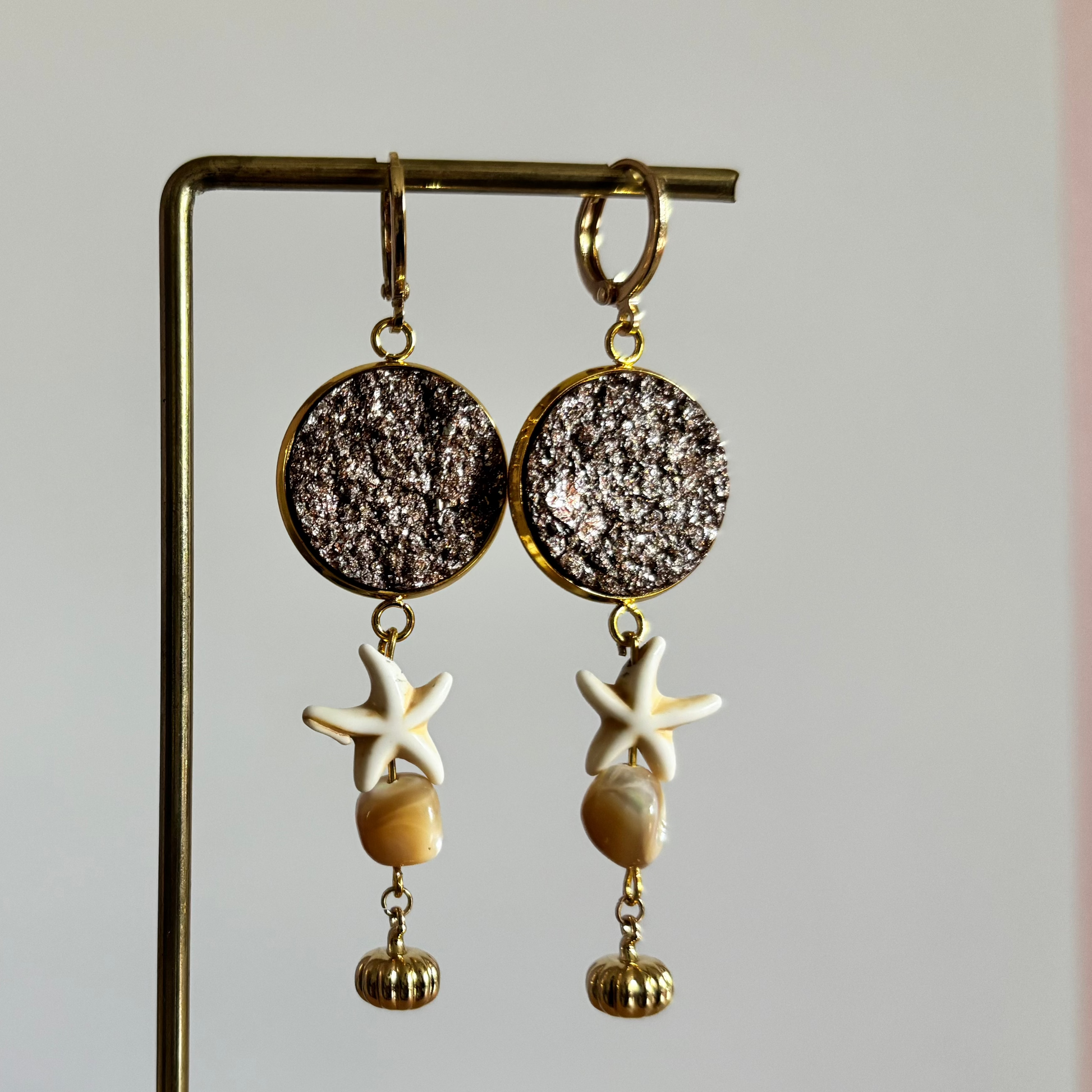 Symmetrische goudkleurige oorbellen Beachy aan een rekje, opgebouwd uit een ronde goudbruine glittersteen, een witte zeester, een beige schelpje en een geribbeld gouden kraaltje.