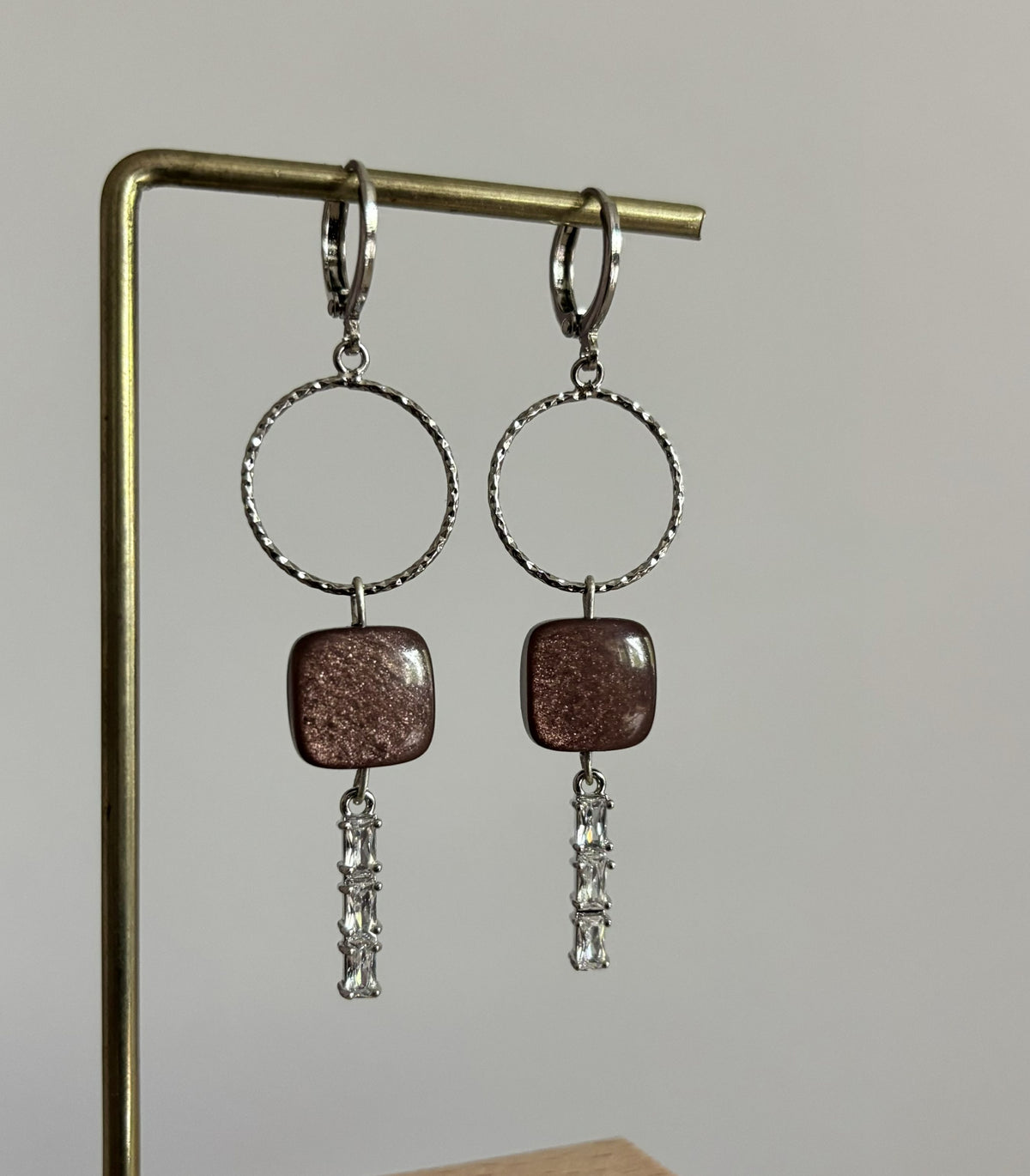 Zilverkleurige oorbellen Bibelot aan een standaard, bestaande uit een bewerkte ronde ring, een vierkante bruine steen met subtiele glitters en drie onder elkaar hangende rechthoekige strass-steentjes.