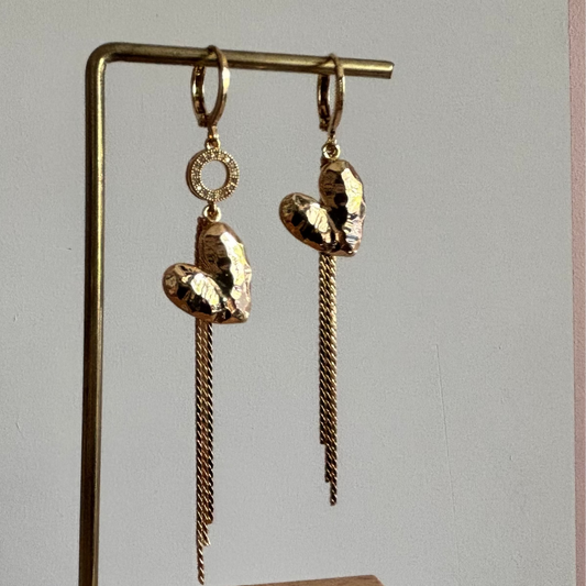 Goudkleurige asymmetrische oorbellen Bibian Goud met gehamerde harten, vallende kettinkjes en een sprankelend strass-ringetje aan één zijde.