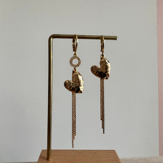 Goudkleurige asymmetrische oorbellen Bibian Goud met gehamerde harten, vallende kettinkjes en een sprankelend strass-ringetje aan één zijde.