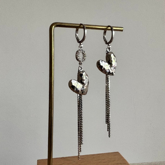 Zilverkleurige asymmetrische oorbellen Bibian met gehamerde harten, vallende kettinkjes en een sprankelend strass-ringetje aan één zijde.