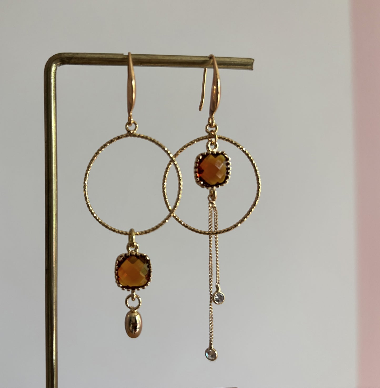 Goudkleurige asymmetrische oorbellen Cara met bewerkte ronde ringen, vierkante terrakleurige kristallen en fijne kettinkjes met zirkonia.