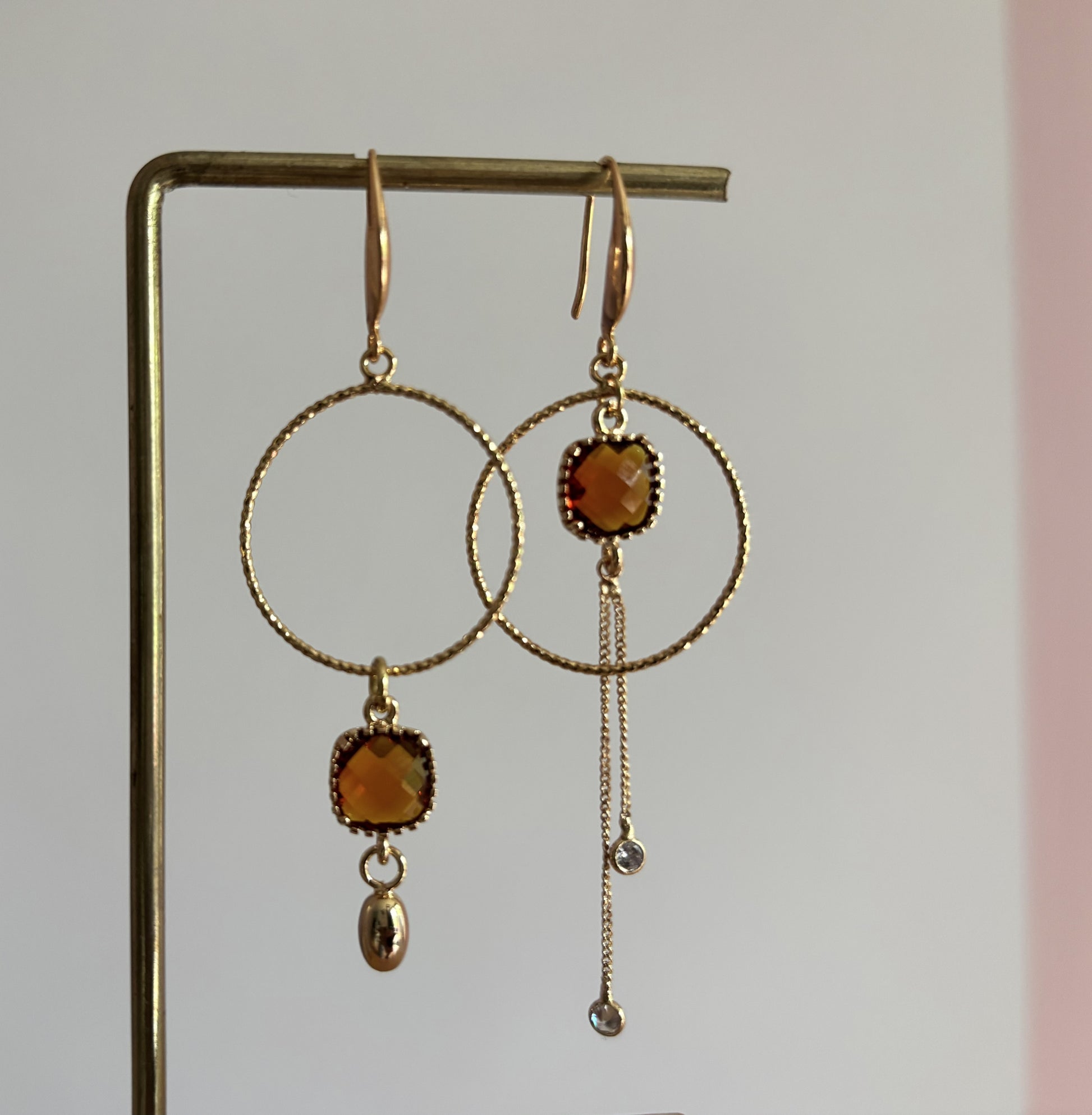 Goudkleurige asymmetrische oorbellen Cara met bewerkte ronde ringen, vierkante terrakleurige kristallen en fijne kettinkjes met zirkonia.