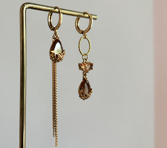 Goudkleurige asymmetrische oorbellen Cathelijne met gefacetteerde amberkleurige druppelstenen, gouden cirkel en een lang neerhangend kettinkje.