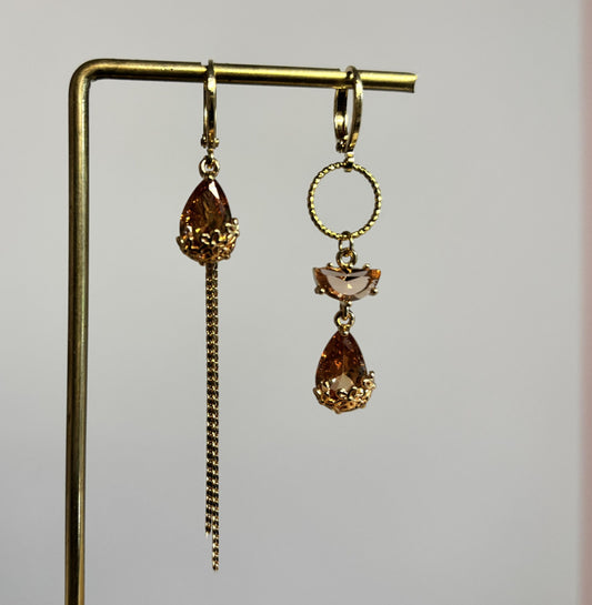 Goudkleurige asymmetrische oorbellen Cathelijne met gefacetteerde amberkleurige druppelstenen, gouden cirkel en een lang neerhangend kettinkje.