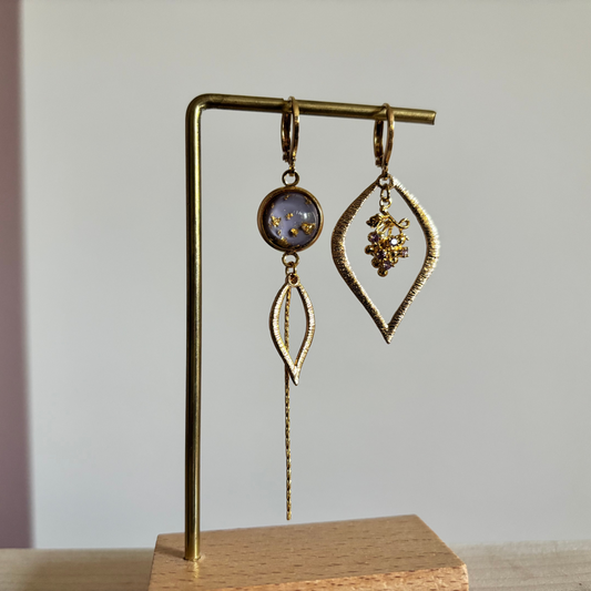 Asymmetrische goudkleurige oorbellen Cathleen aan een rekje. Links een lila ronde steen met goudspikkels, een bladvormige hanger en een lang kettinkje; rechts een grote open ruitvorm met een klein paars-gouden druiventrosje erin.