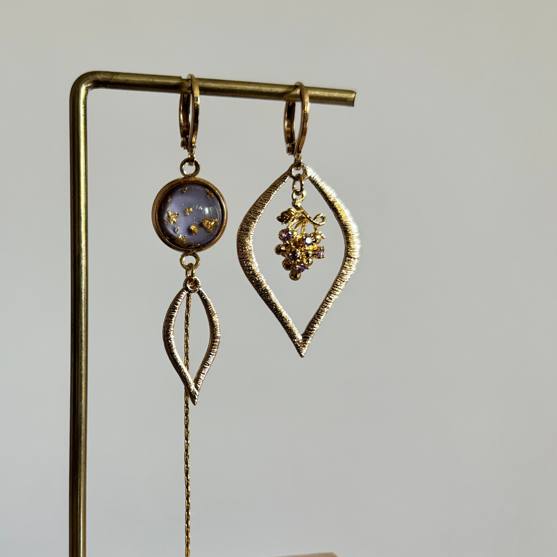 Asymmetrische goudkleurige oorbellen Cathleen aan een rekje. Links een lila ronde steen met goudspikkels, een bladvormige hanger en een lang kettinkje; rechts een grote open ruitvorm met een klein paars-gouden druiventrosje erin.