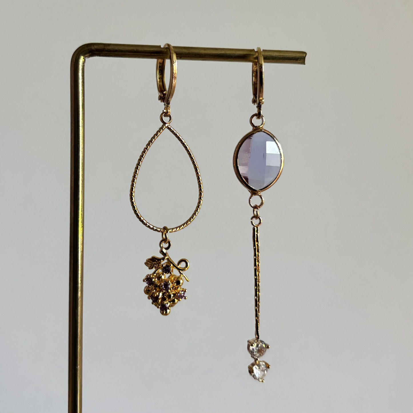 Set asymmetrische goudkleurige oorbellen aan een display. Links een ronde lila facetgeslepen steen met daaronder een fijn goudkleurig staafje en twee transparante steentjes. Rechts een grote opengewerkte druppelvorm met onderaan een bedel in de vorm van een druiventros.